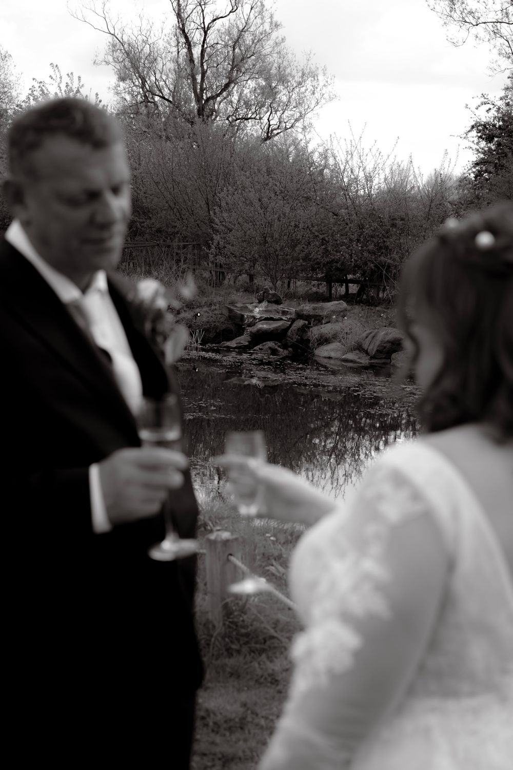 A black and white wedding photo of a couple toasting with champagne glasses by a serene pond.