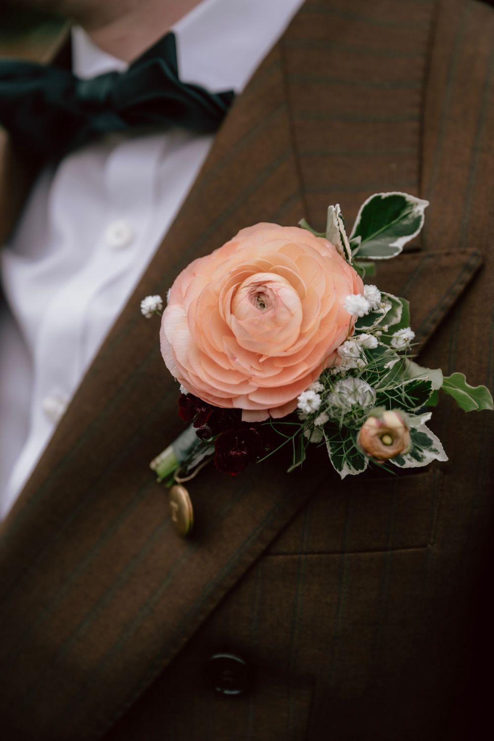 A delicate peach ranunculus boutonniere with greenery and baby's breath pinned to a brown suit lapel.