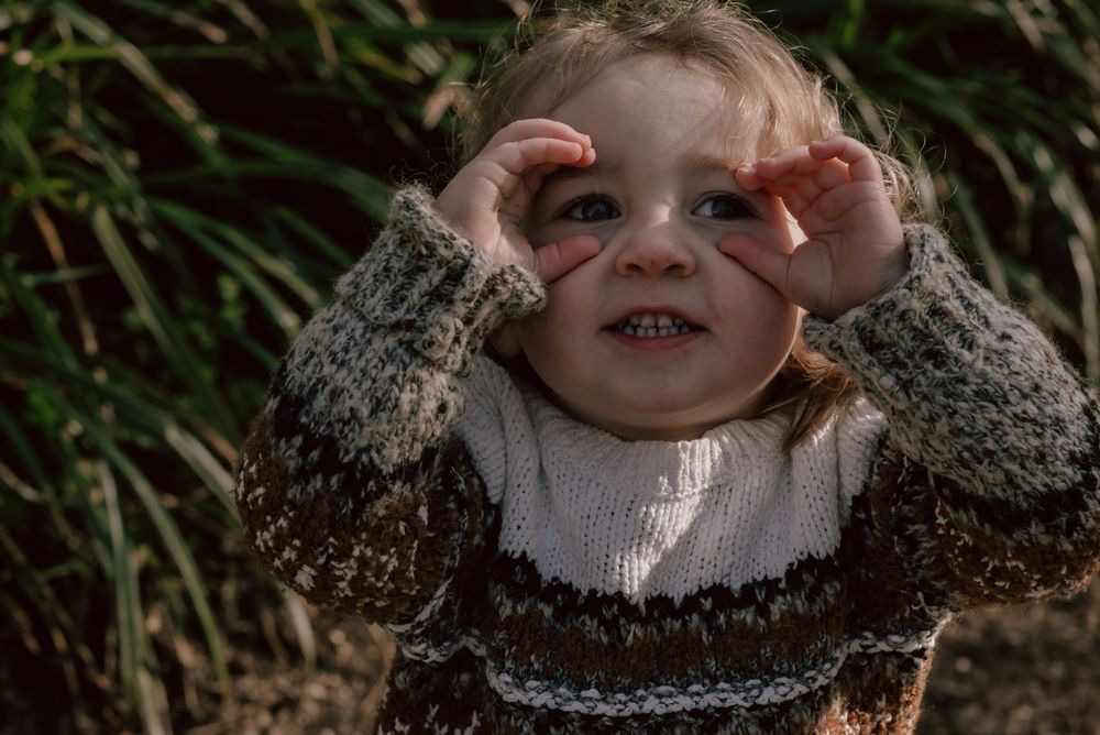 A small child wearing a knit sweater smiles while playfully covering their eyes against a natural backdrop of foliage.
