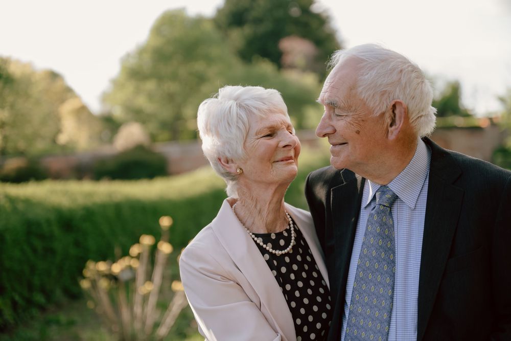 Happy elderly couple smiling at each other outdoors in a lush garden, dressed in elegant formal attire during wedding anniversary photos