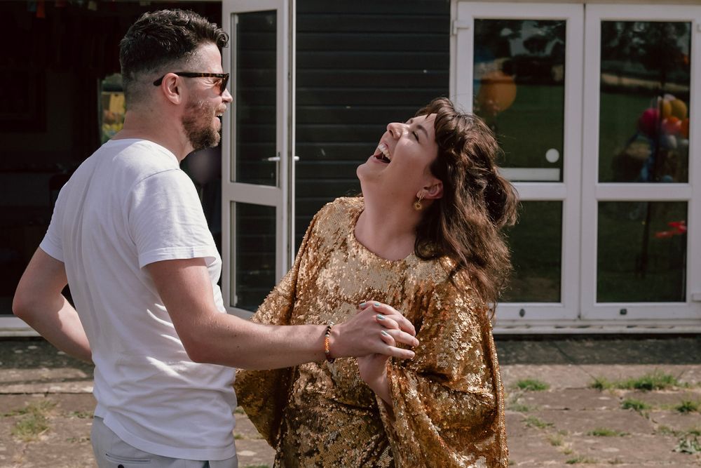 A man and woman laughing and dancing together outdoors on a sunny day celebrating a 40th birthday wearing a gold sequin kaftan