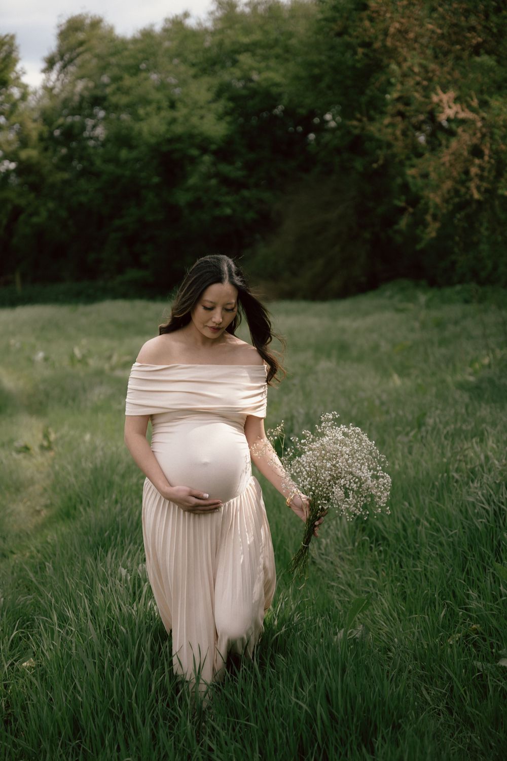 Maternity photo shoot in a serene meadow with tall grass, featuring someone in a flowing blush-colored gown at sunset.