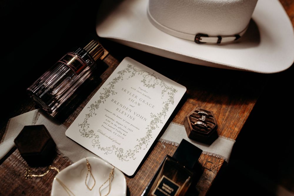 Wedding flat lay featuring invitation, white cowboy hat, perfume bottles, and accessories on wood surface.