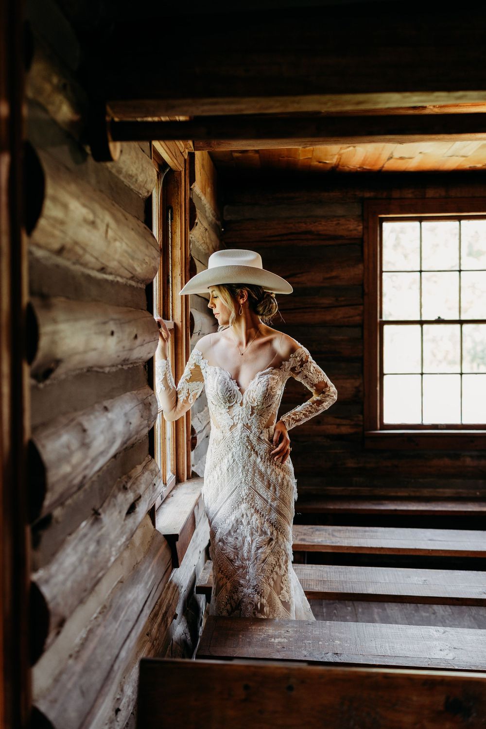 Bride in lace gown and white cowboy hat poses in rustic log cabin stairway for wedding portraits.