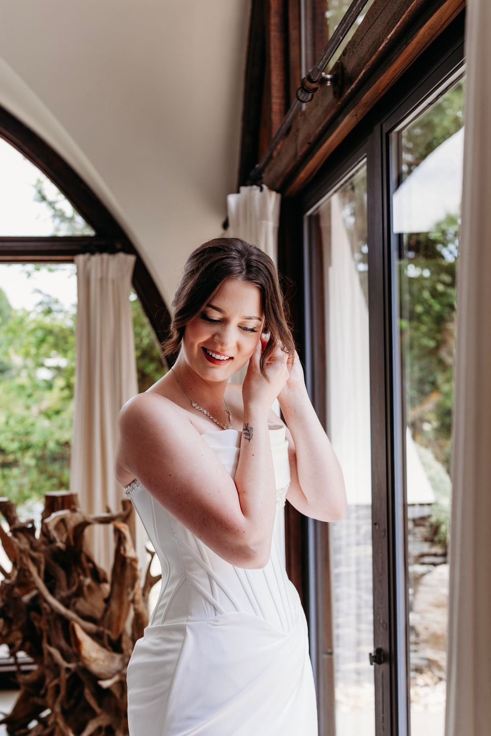 Smiling bride in white strapless gown adjusts earring near large arched windows at Waterstone Venue in Johnson City, TN.