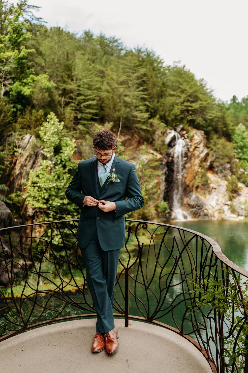 Dapper groom in green suit buttoning jacket on balcony overlooking quarry lake and waterfall backdrop at Waterstone Venue in Johnson City.