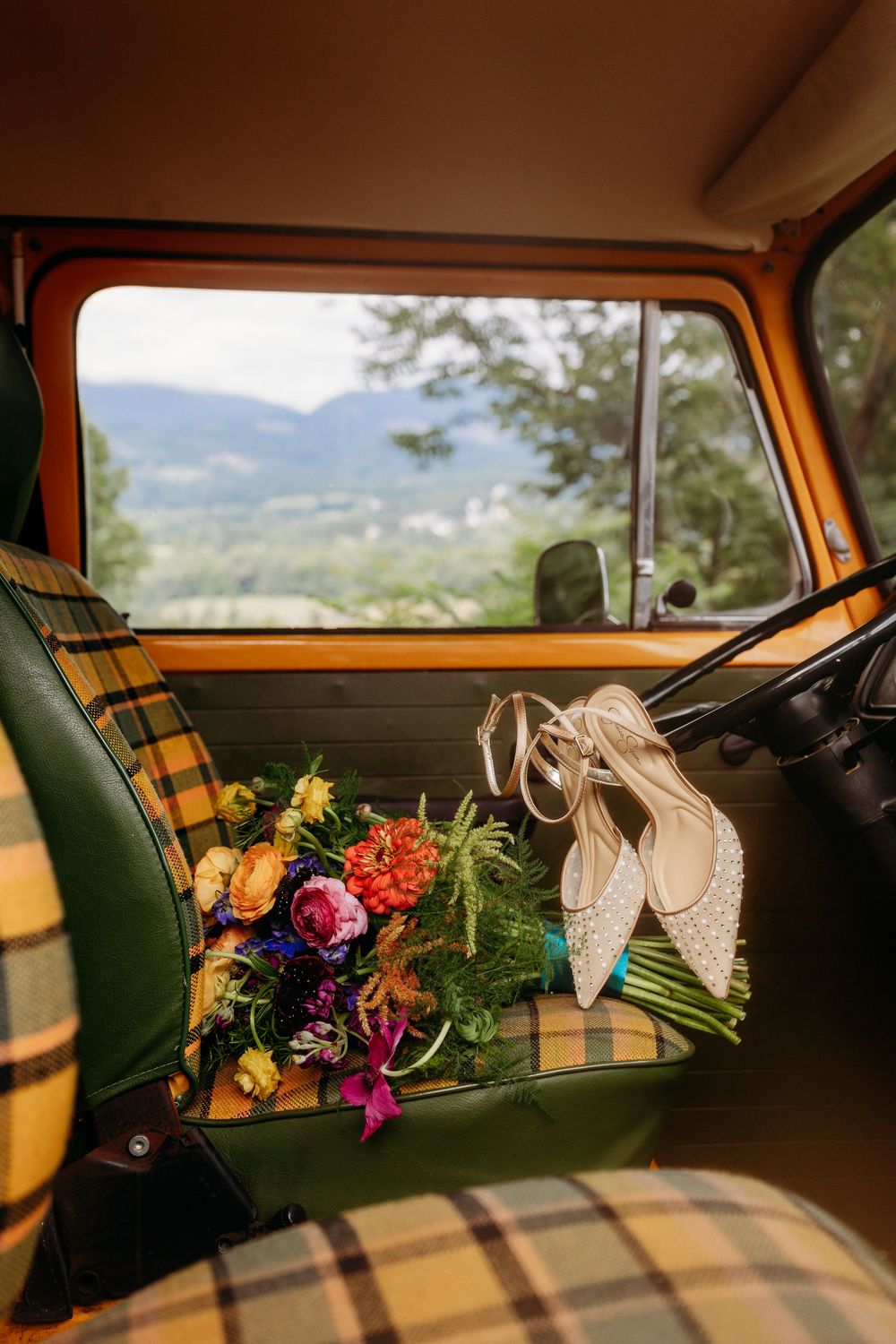 Colorful wedding bouquet and bridal heels on plaid seat of vintage VW bus with mountain view.