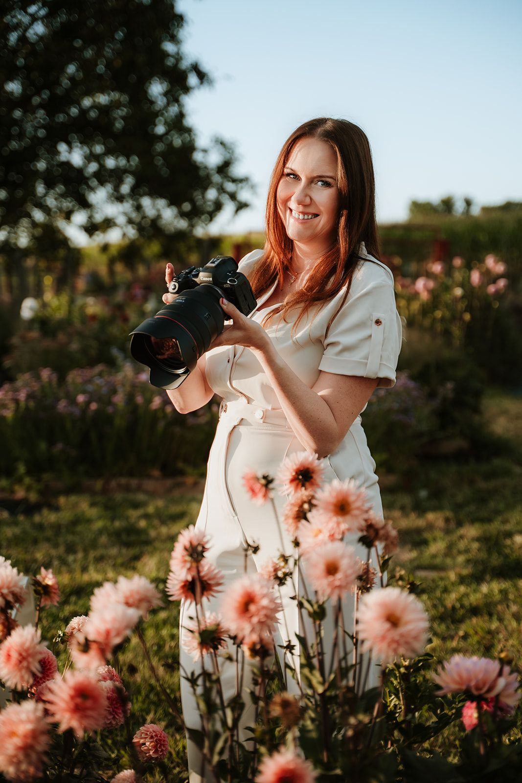 A photographer smiling while holding a professional camera and sitting on a wooden stool against a white background.
