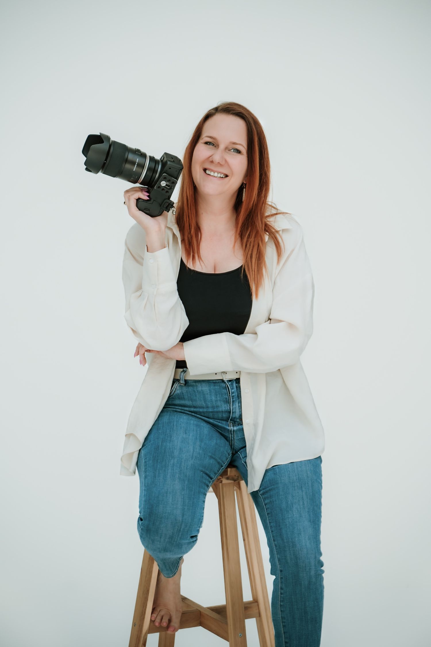 A photographer smiling while holding a professional camera and sitting on a wooden stool against a white background.