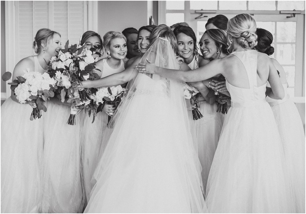 Wedding party in elegant white dresses huddle together in joyful embrace during celebration moment.