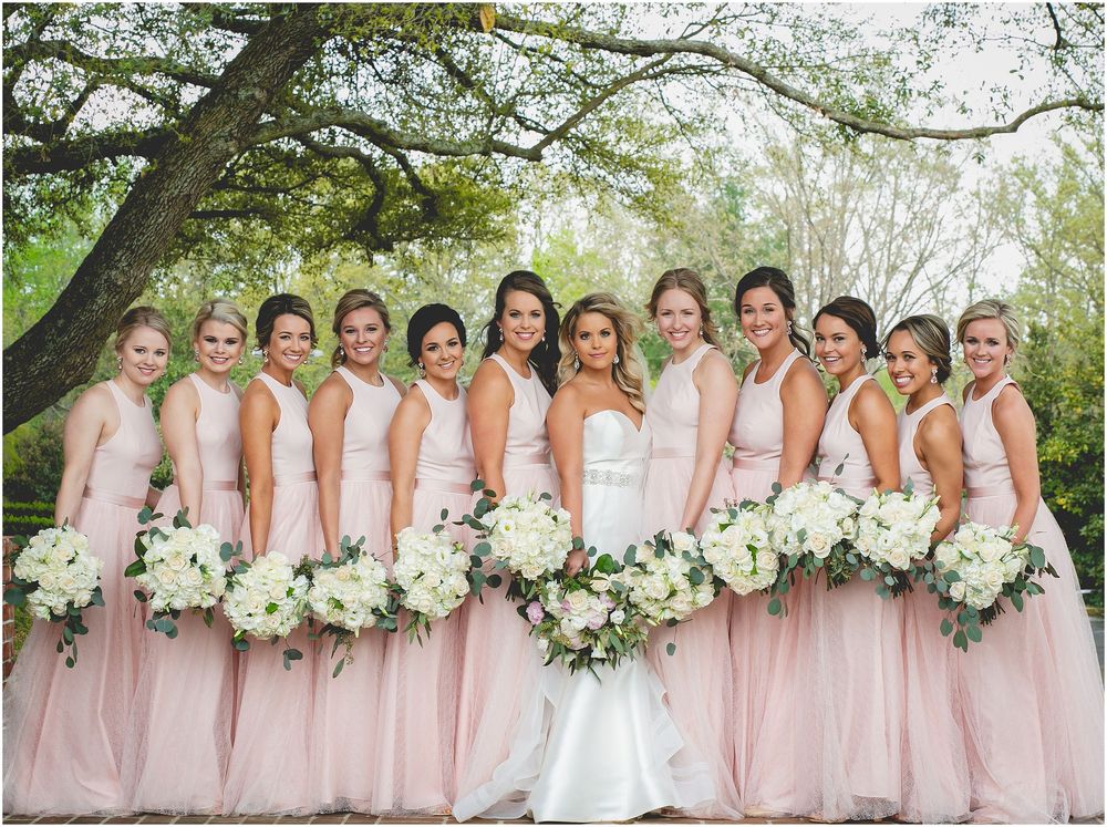Wedding party in blush pink bridesmaid dresses poses with white floral bouquets in an outdoor garden setting.