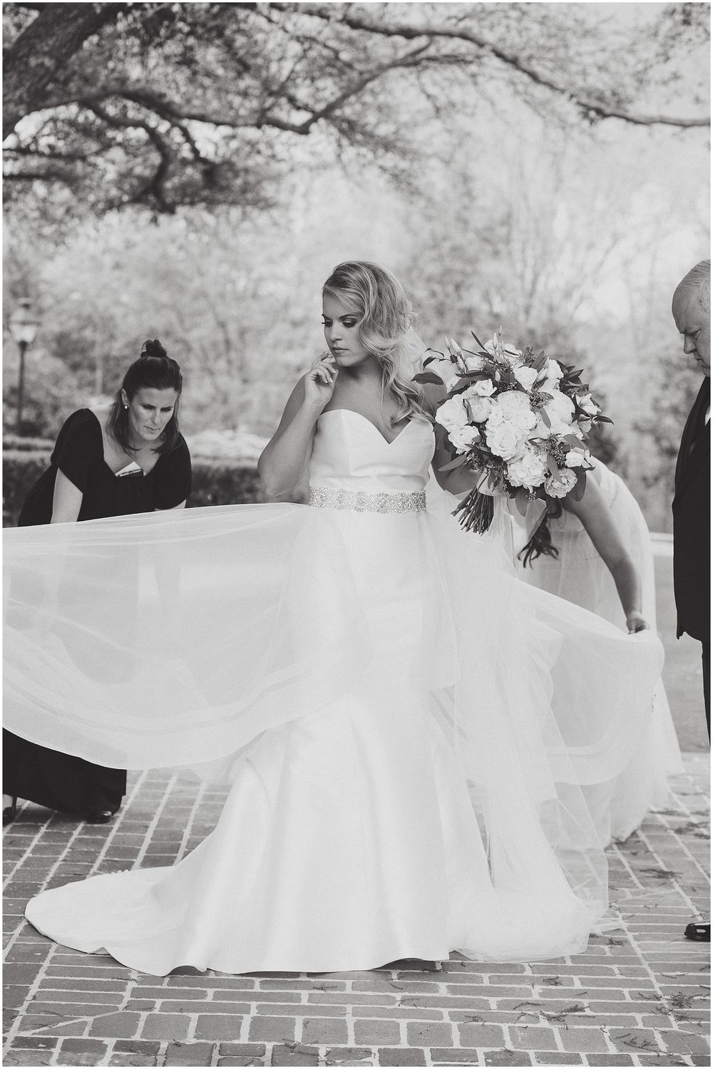 A row of bridesmaids in white dresses kneel on brick pavement holding bouquets during an outdoor wedding photo.
