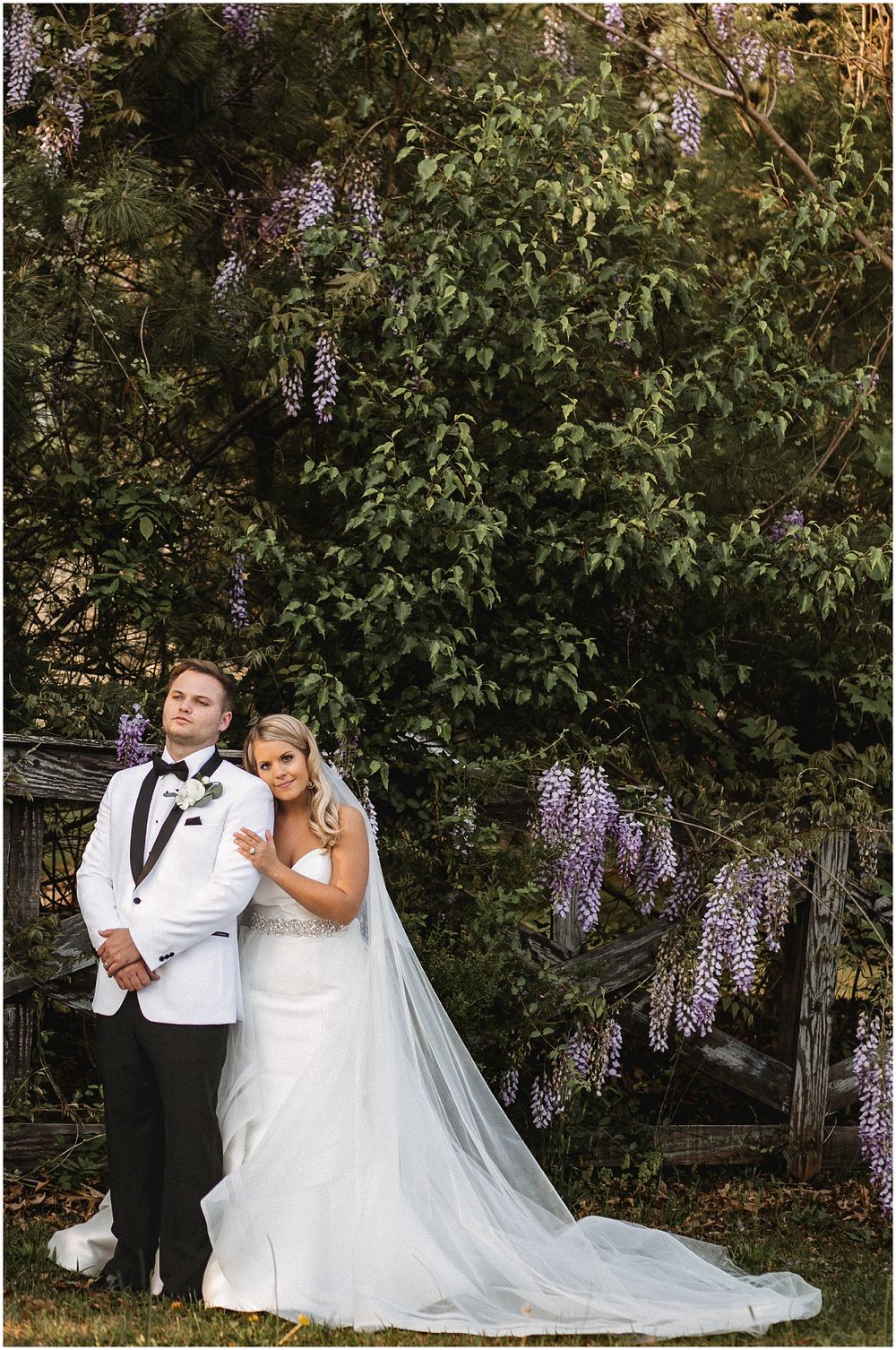 A couple in wedding attire poses under blooming wisteria flowers in a garden setting.