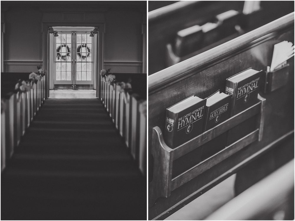 Black and white image of empty church pews with ornate wooden details and stained glass windows in background.