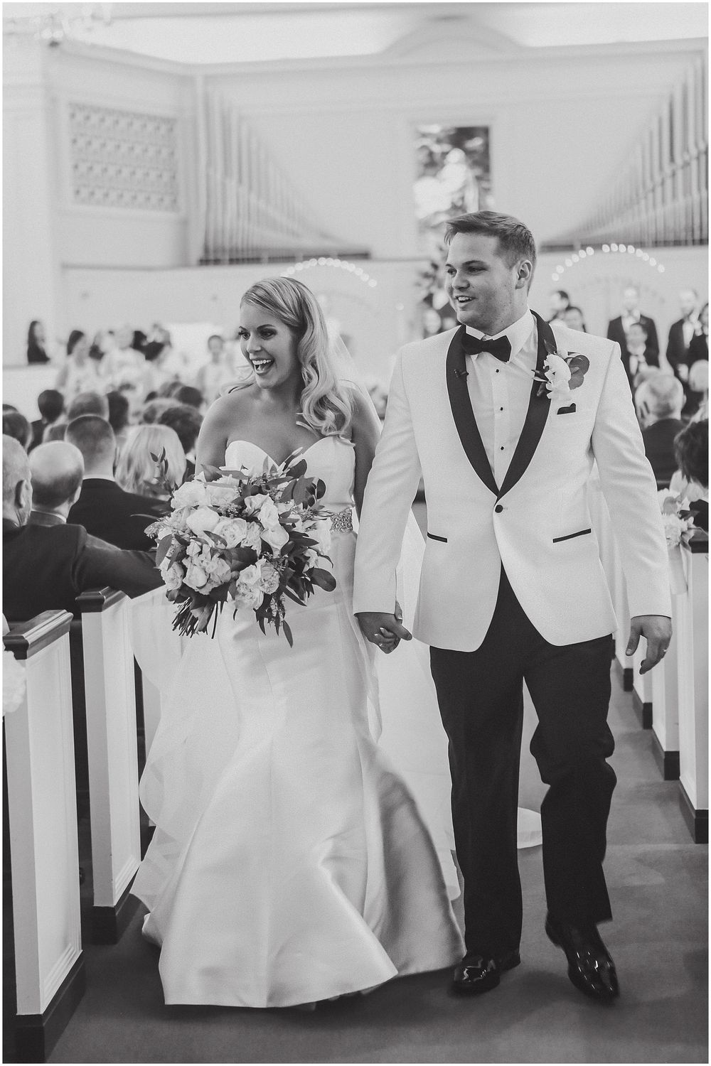 Wedding party members in white and black formal attire walk together while holding bouquets in an elegant venue.