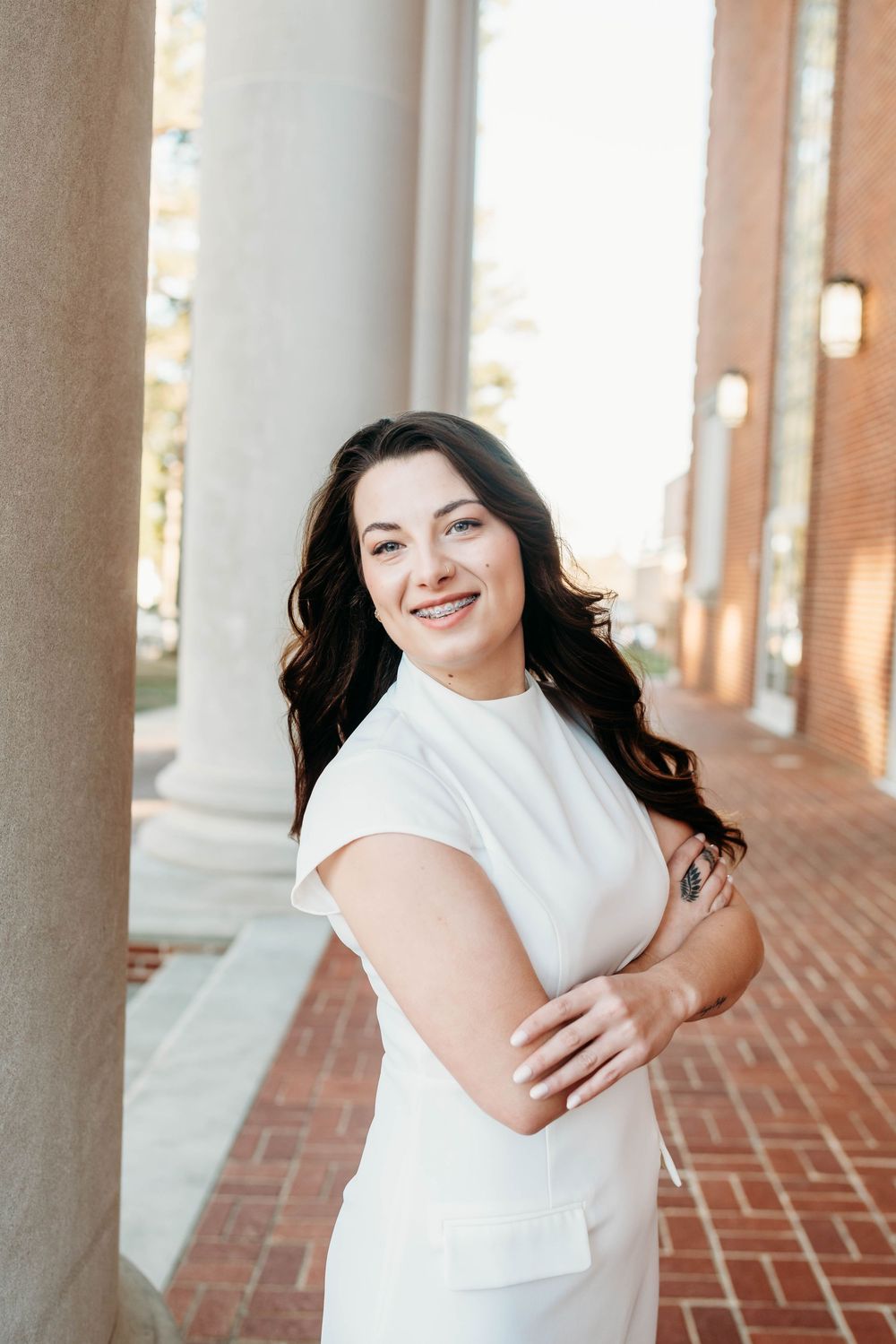 A smiling woman in a white dress poses confidently with arms crossed between brick columns in an outdoor setting.