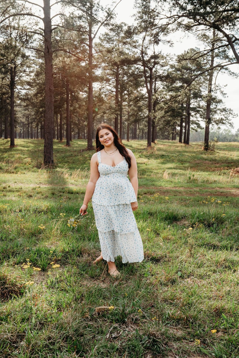 A smiling woman in a white floral midi dress stands in a sunlit forest clearing with tall pine trees.