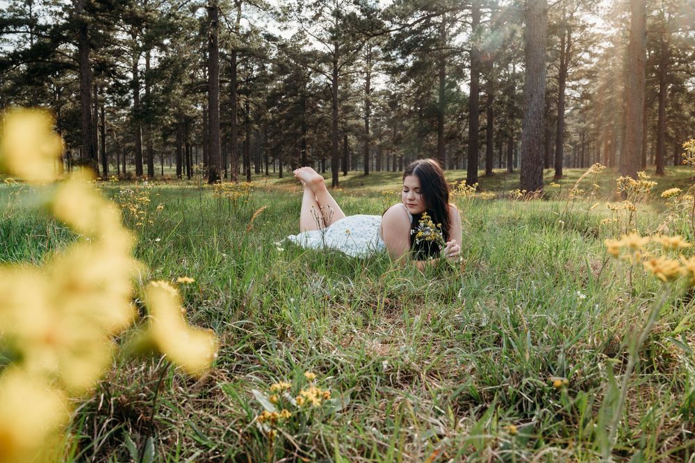 Woman in white dress lying in sunny forest meadow surrounded by yellow wildflowers and tall pine trees.