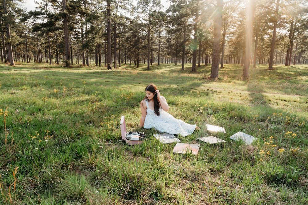 Woman in white dress sitting on blanket surrounded by books in sunlit forest clearing during golden hour.