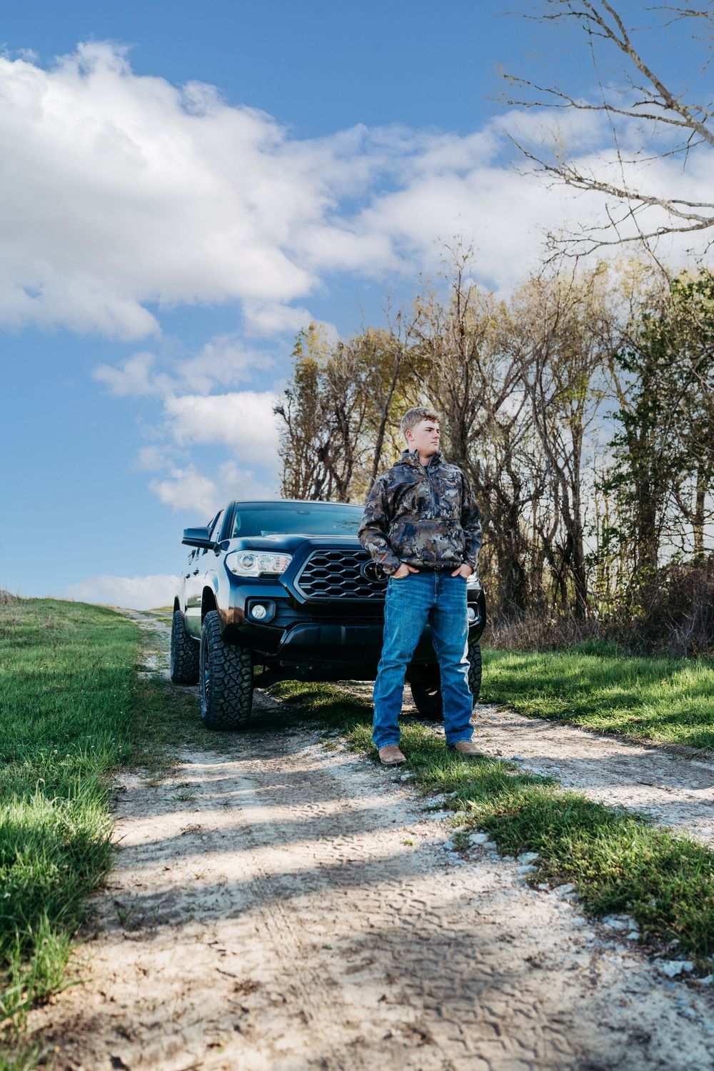 Man in camo jacket and jeans leaning against a black pickup truck on a dirt road surrounded by green fields and trees.