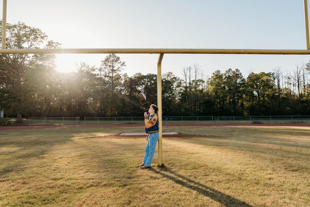 Person leaning against a football field goal post during golden hour sunset with trees in background.