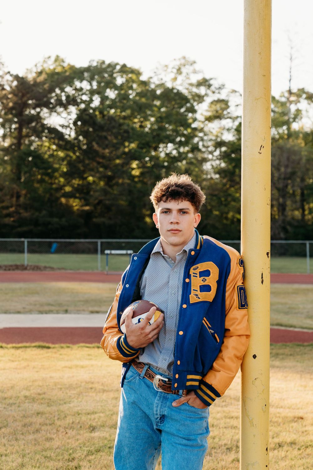 Teen boy in blue and gold varsity jacket holding football, leaning on goalpost at high school field.
