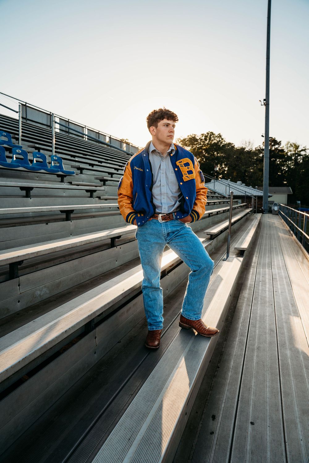 Young man in varsity jacket and jeans poses on bleachers at a football stadium during golden hour.