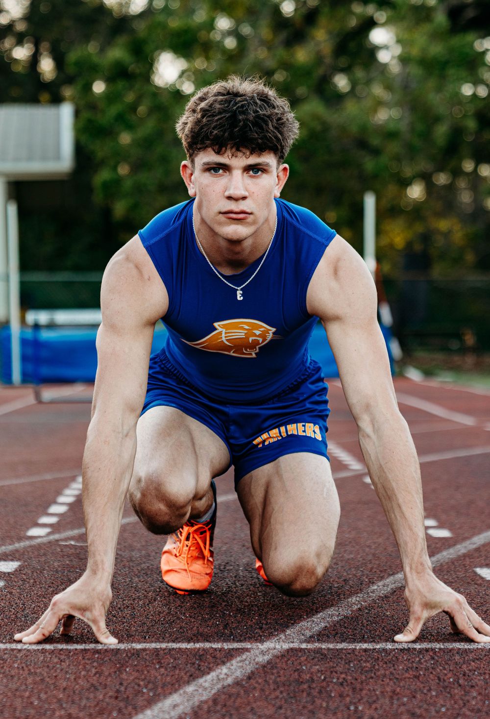 A young male track athlete in a blue Panthers uniform crouches in a starting position on a red running track.