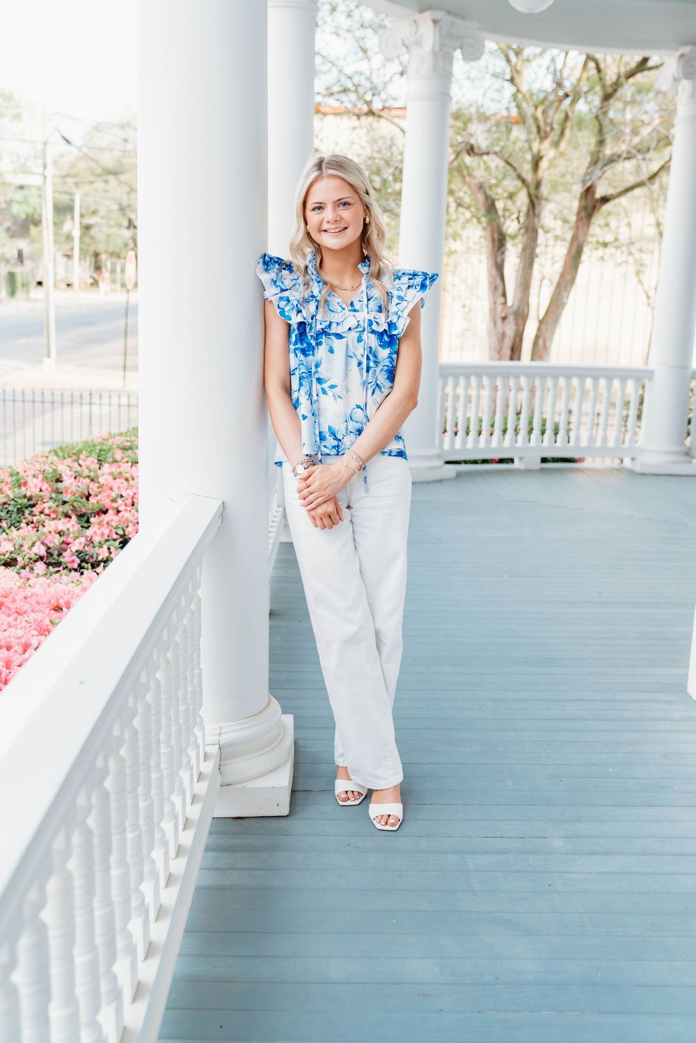 Blonde woman in blue floral top and white pants posing on classic Southern porch with columns.