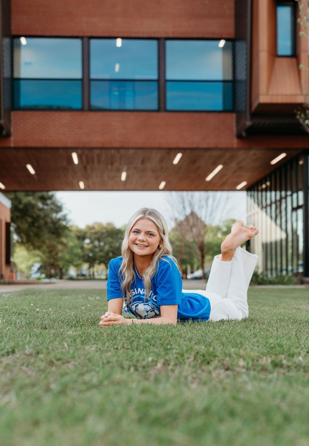 Smiling blonde woman in blue college shirt lying on green grass in front of modern brick building on sunny day.