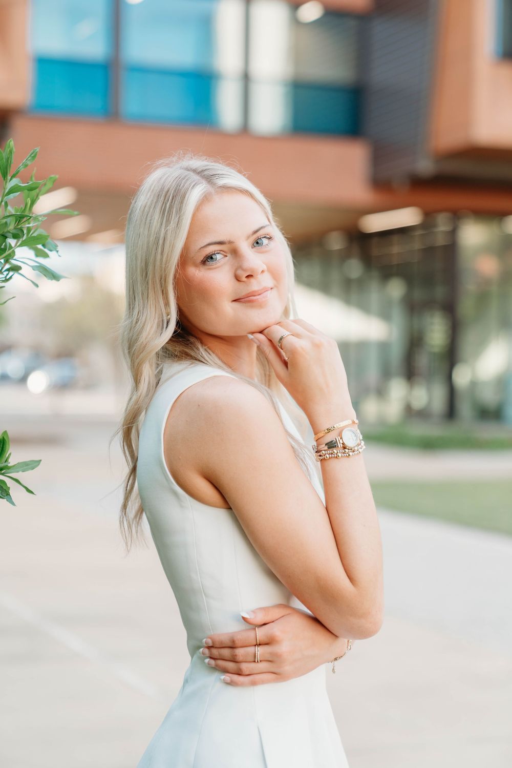 Young blonde woman in white sleeveless dress posing outdoors near modern building, smiling with hand on chin.