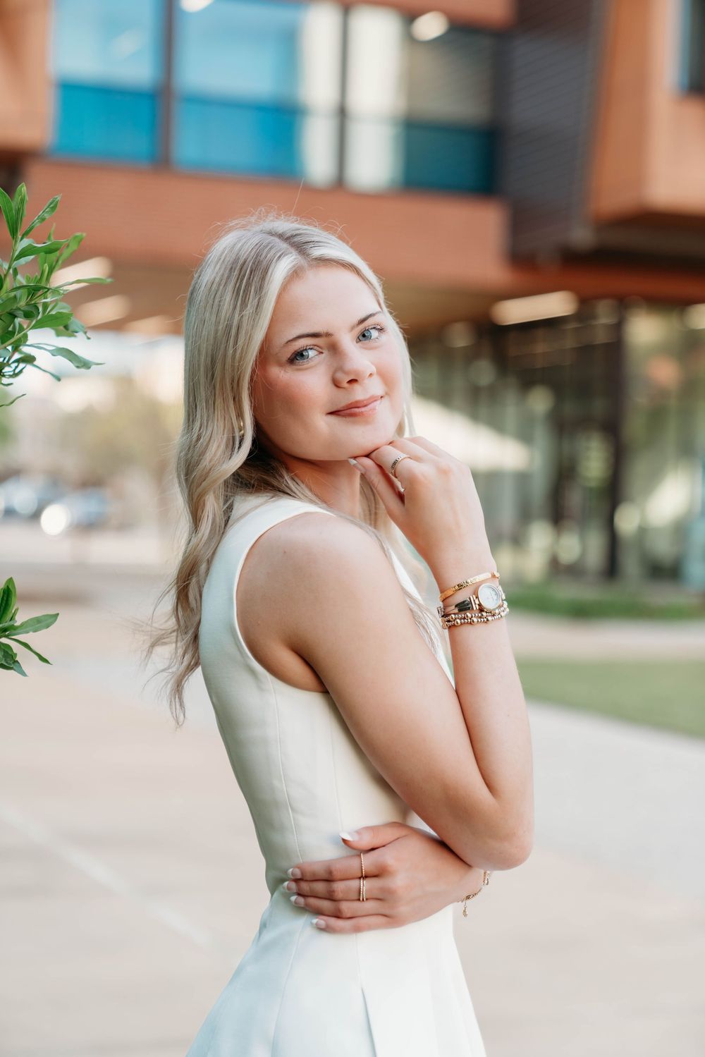 Young blonde woman in white dress posing outdoors near modern building, smiling with hand on chin.