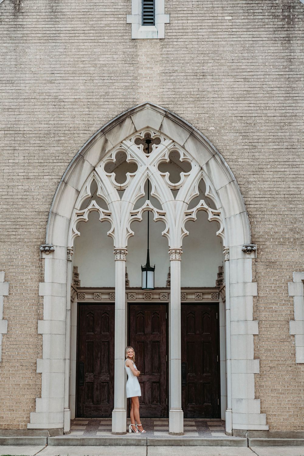 Blonde woman in white dress standing under Gothic arch entrance of historic brick church.
