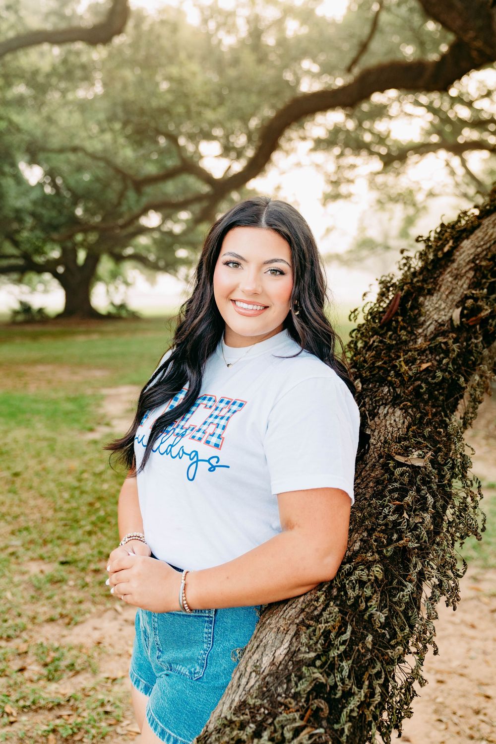 Young woman with dark hair smiling in a white graphic tee and denim shorts, leaning against a moss-covered oak tree.