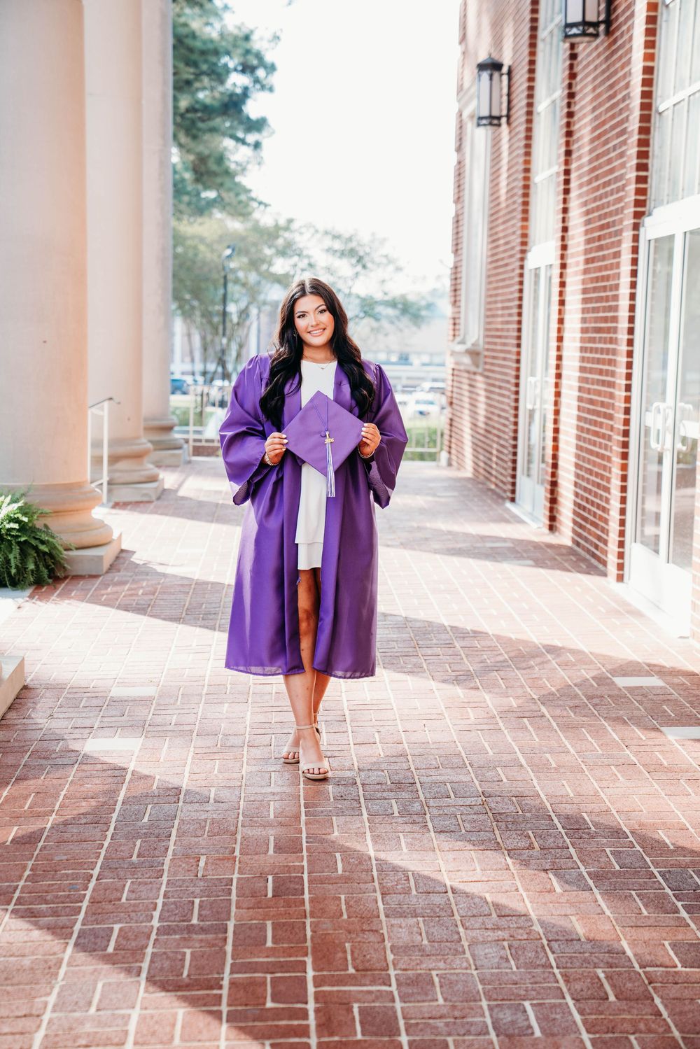 Smiling young woman in a purple graduation gown and cap posing on a brick walkway outside a building.