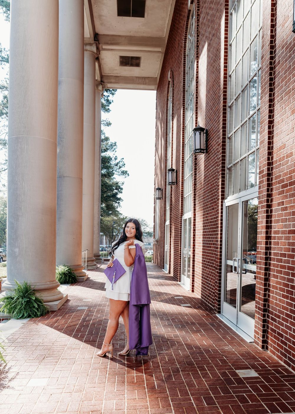 A graduate poses in a purple cap and gown on a brick walkway beside white columns of a university building.