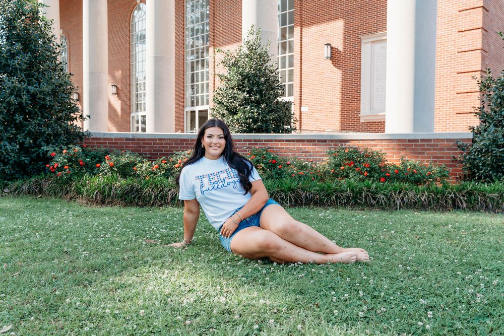 Young woman in a graphic tee and denim shorts sitting on green grass in front of a brick building.