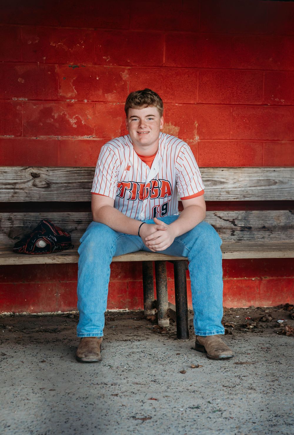 Young baseball player in a Tioga jersey sits smiling on a dugout bench with a glove nearby.