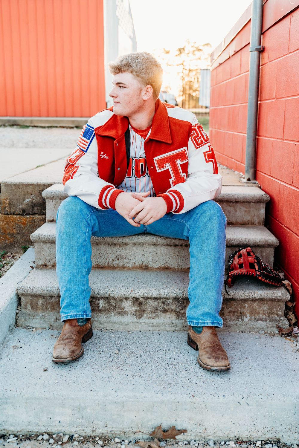 Young man in a red varsity letterman jacket and jeans sitting on concrete steps near a brick wall.
