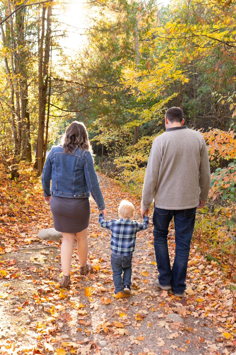 Family walking together on autumn trail covered in fallen orange and yellow leaves in forest setting.