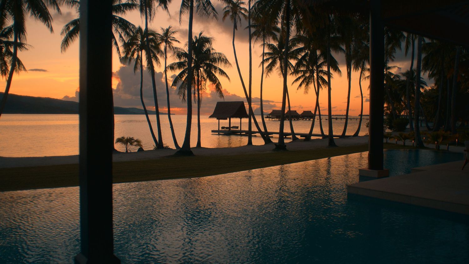 A silhouetted figure admires the ocean view from an empty beachfront restaurant with bar seating at sunset.