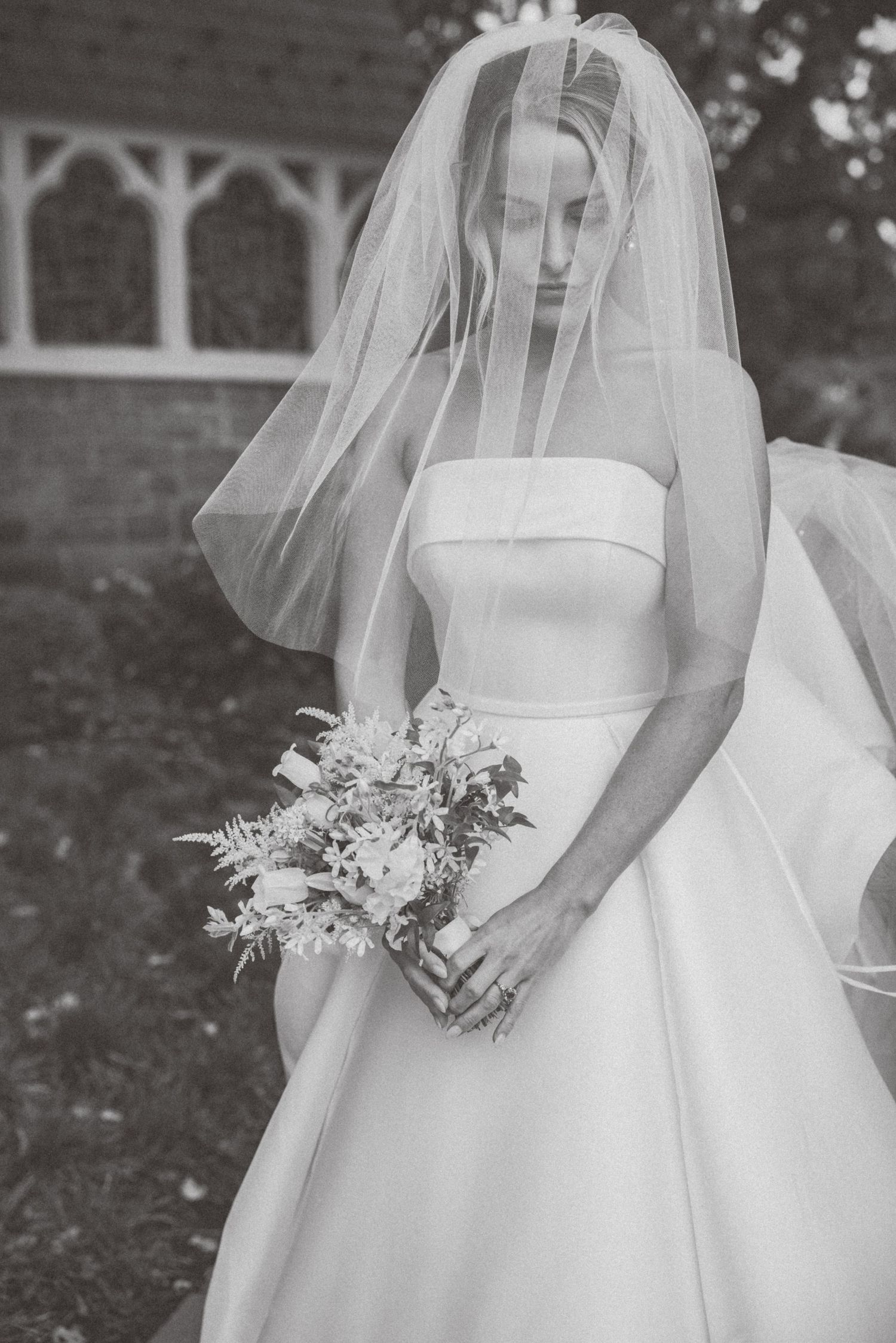 Black and white portrait of bride in strapless gown and veil holding bouquet outdoors.
