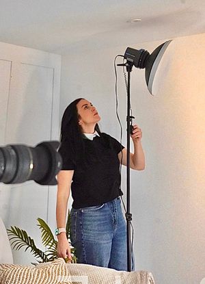 Person in black top and jeans poses against white wall while holding camera equipment and lighting setup.