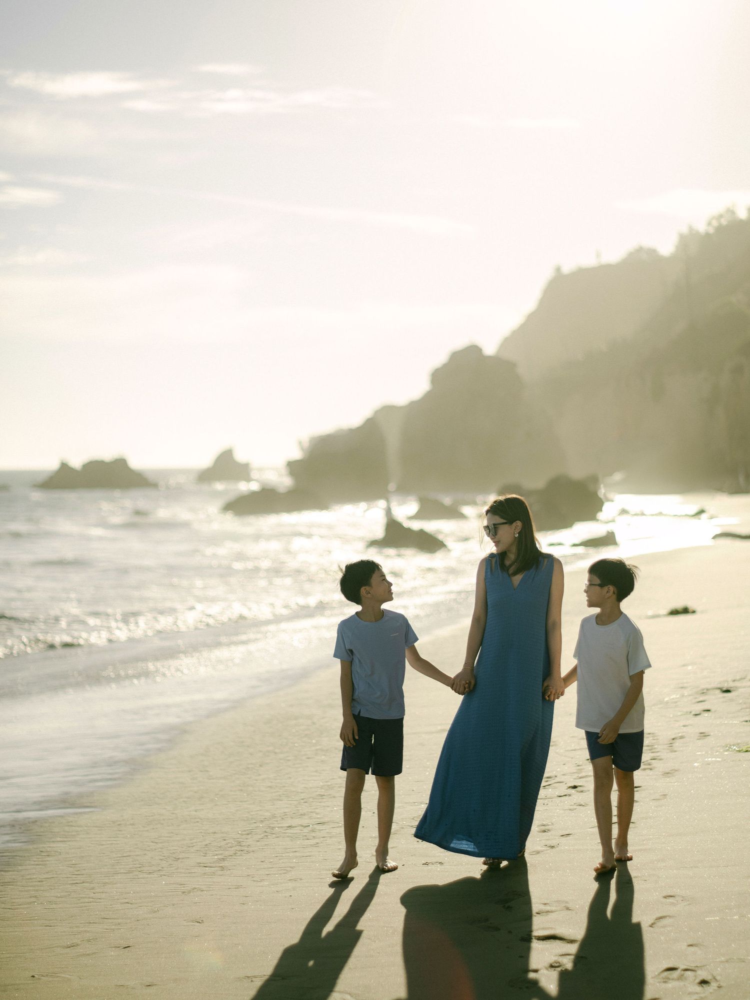 A woman in a blue dress holds hands with two children on a sunlit beach with rocky cliffs in the background.