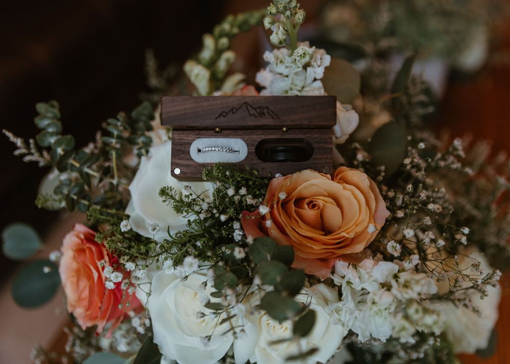 Wooden ring box engraved with mountains resting on a colorful wedding bouquet of orange, coral, and white flowers.