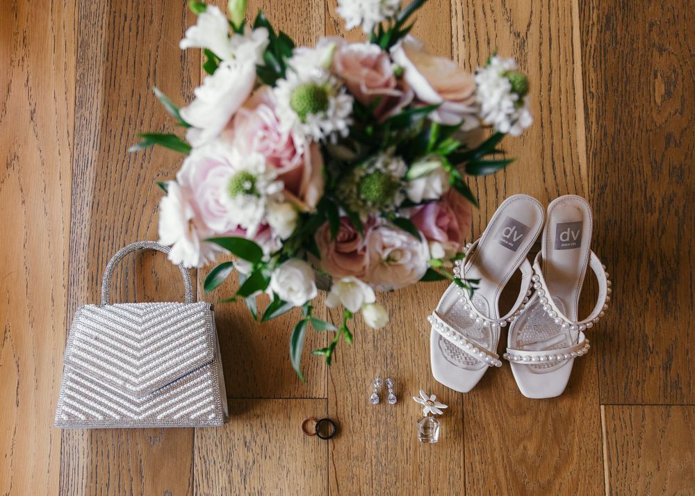 Bridal flat lay featuring a striped clutch, white heeled sandals, floral bouquet, and delicate jewelry on wood floor.