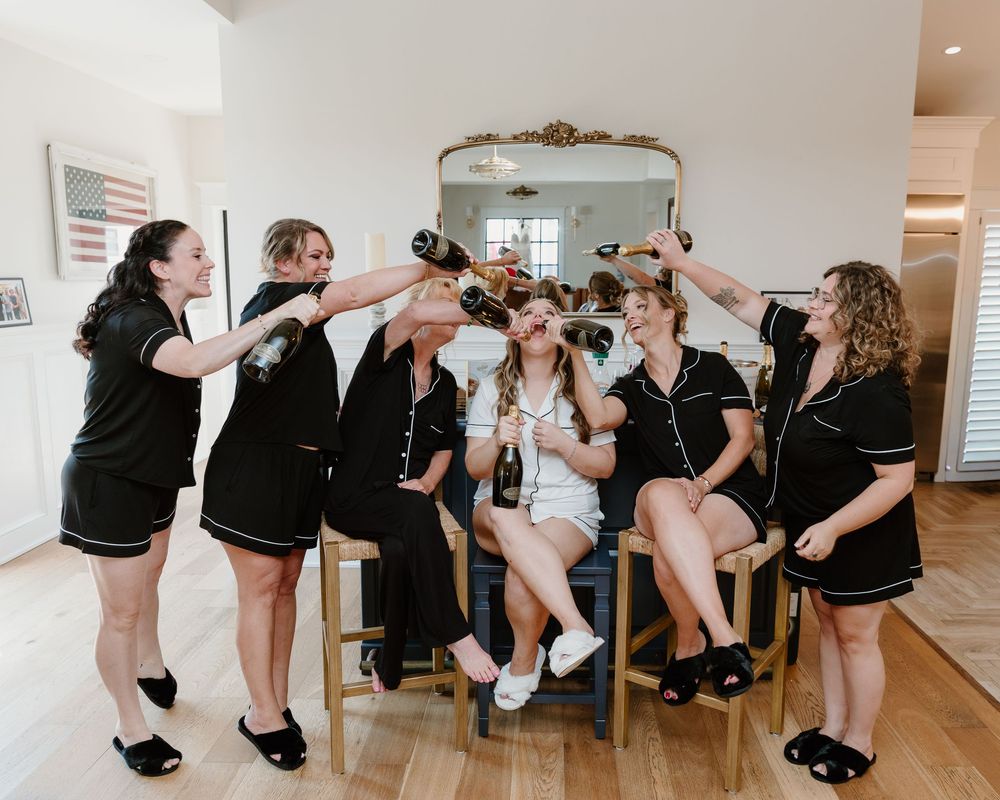 Bridesmaids in black pajamas cheerfully styling the bride's hair and makeup during wedding morning preparations indoors.