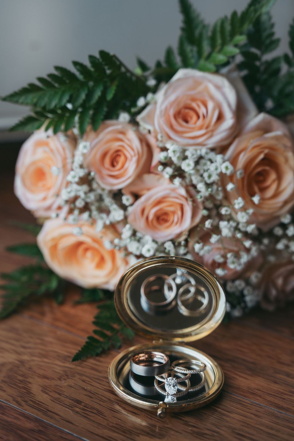Wedding rings displayed in a gold compact mirror beside a bouquet of peach roses and baby's breath on a wooden table.