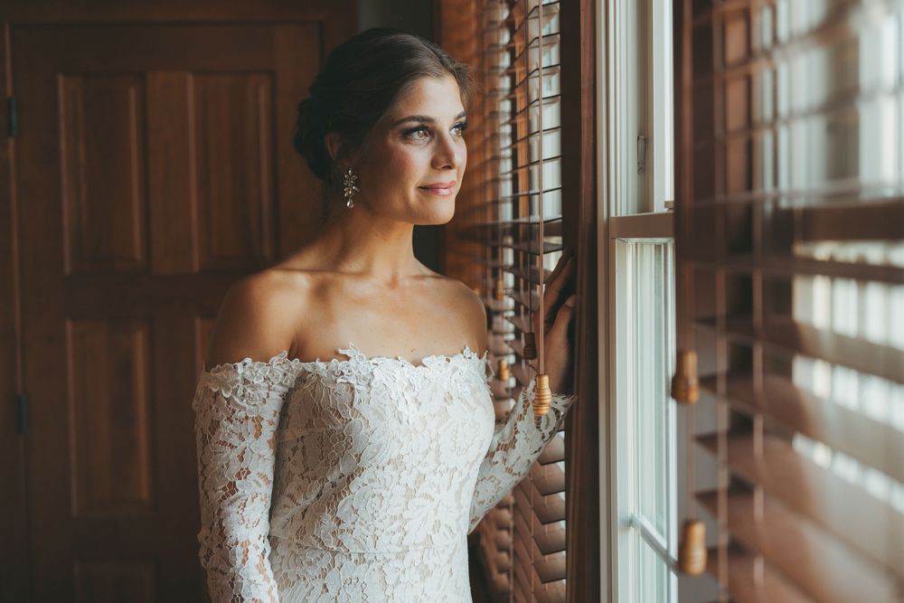 Elegant bride in lace off-shoulder gown gazing through wooden blinds.