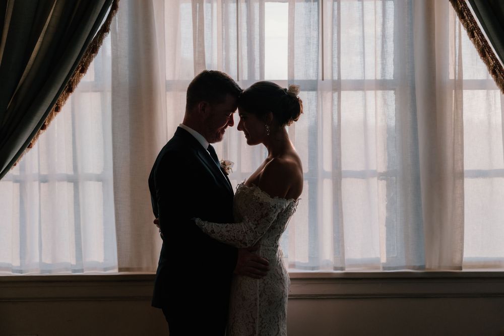 Silhouette of bride and groom embracing near bright window indoors.